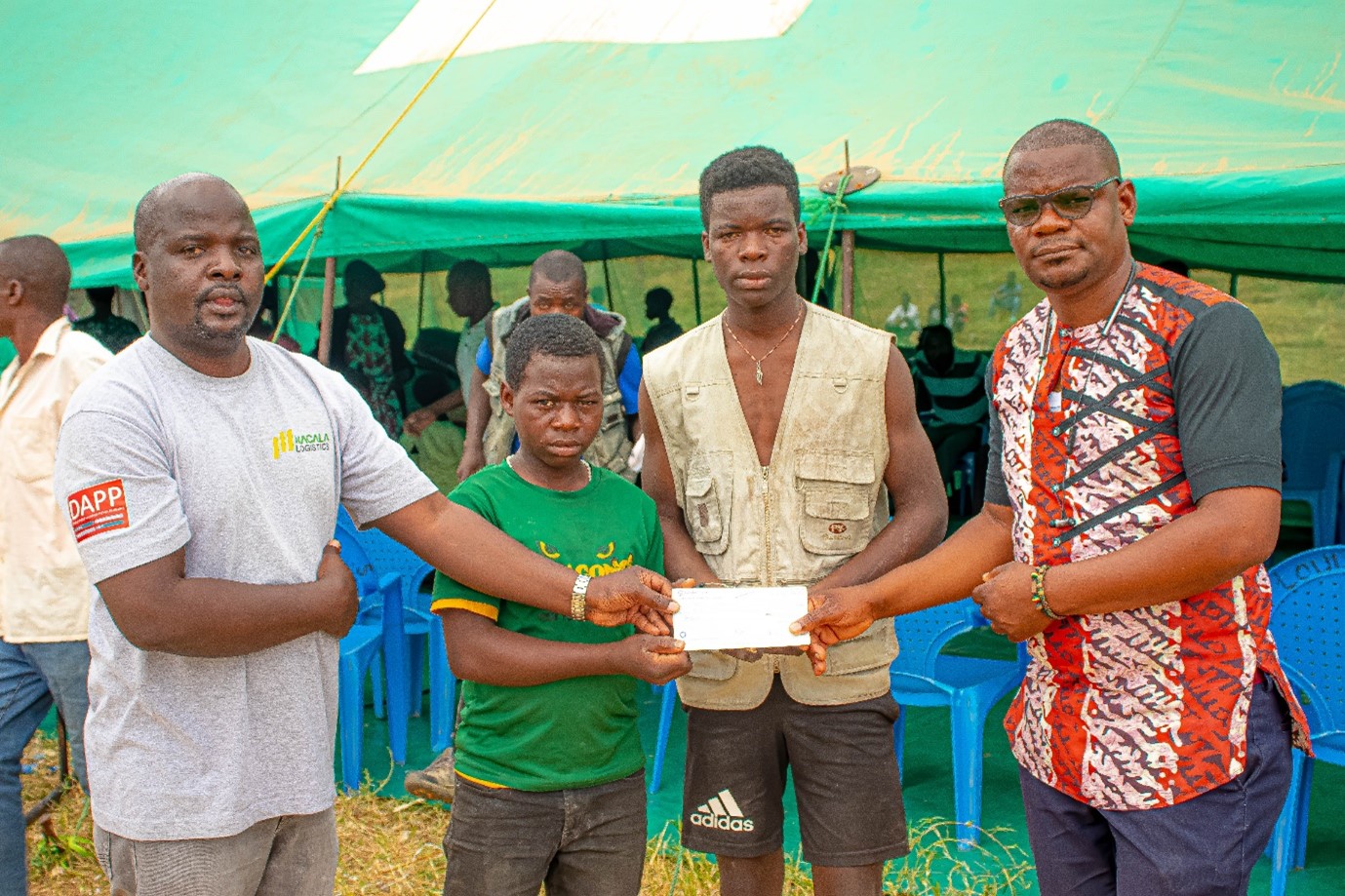 Robert in green shirt and his elder brother Witness in brown vest receiving a cheque from DAPP Official in presence of District Official